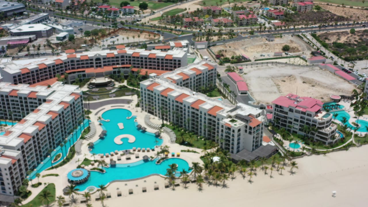 Opening overhead frame in Los Cabos, Mexico shows white resort buildings around turquoise pools with palm-lined paths, nearby roads, sandy open lots, and no visible people.