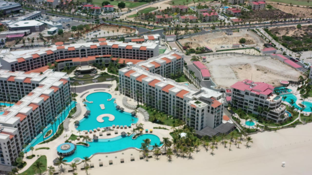 Second overhead frame in Los Cabos, Mexico centers the curved resort buildings, bright pool courtyard, roads, a broad sandy construction area, and no visible people.
