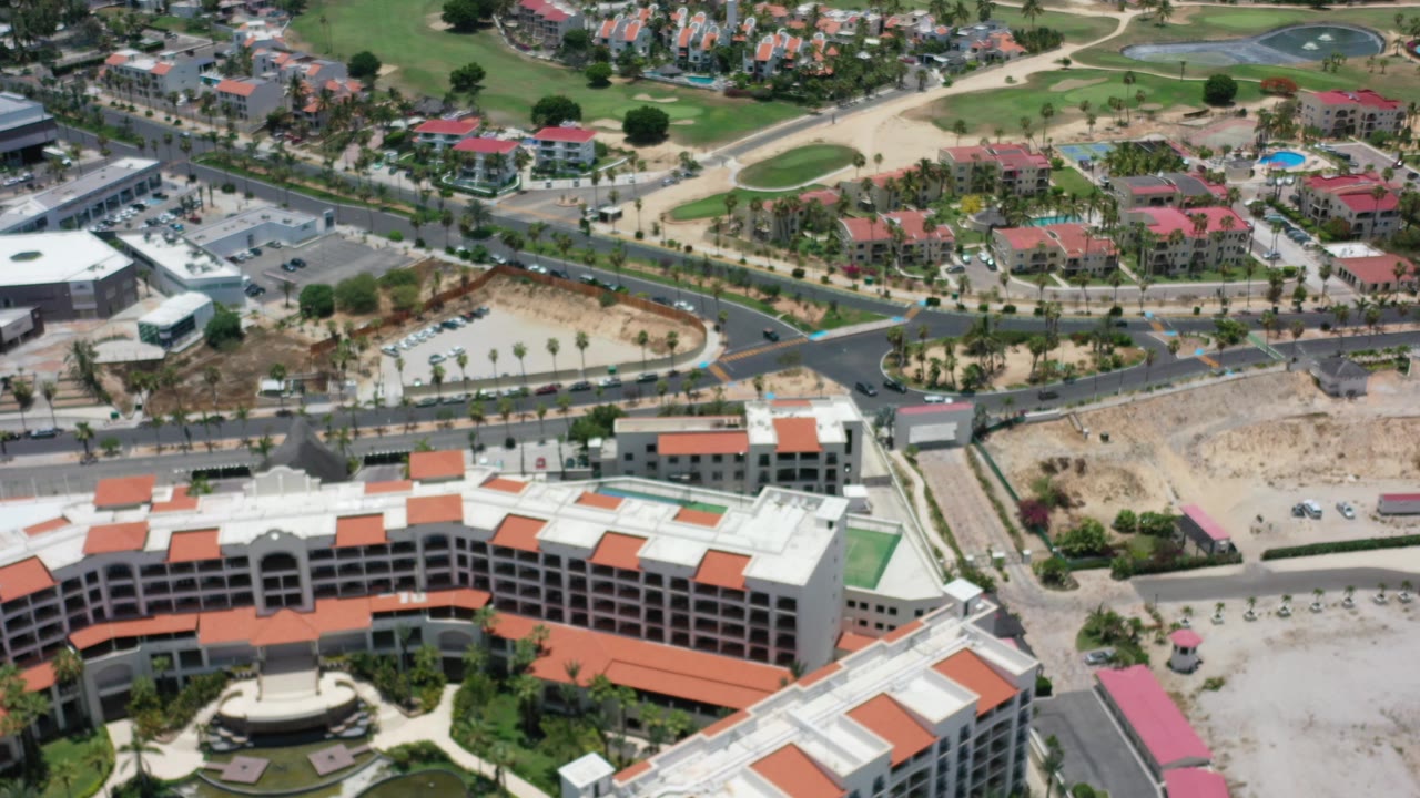 High aerial view over Los Cabos, Mexico, with a white hotel at the bottom, curved roads, small cars, green lawns, red-roofed buildings, and no visible people.