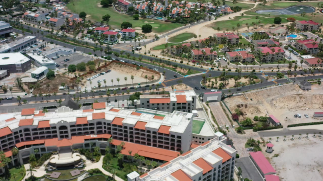 Bird's-eye frame in Los Cabos, Mexico, showing the hotel roofline lower in frame, intersecting roads, parked and moving cars, landscaped areas, and no visible people.