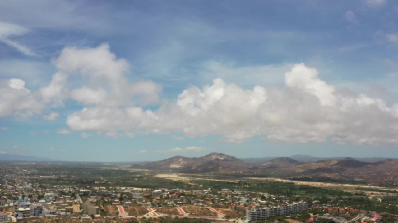 Opening frame of Los Cabos, Mexico from high above, showing low city blocks, green areas, rugged hills, and a bright cloud-filled sky with no visible people.