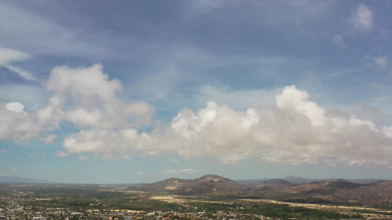 A broad hazy sky holds bright white cloud masses above brown hills and a thin strip of Los Cabos terrain, with no visible people.