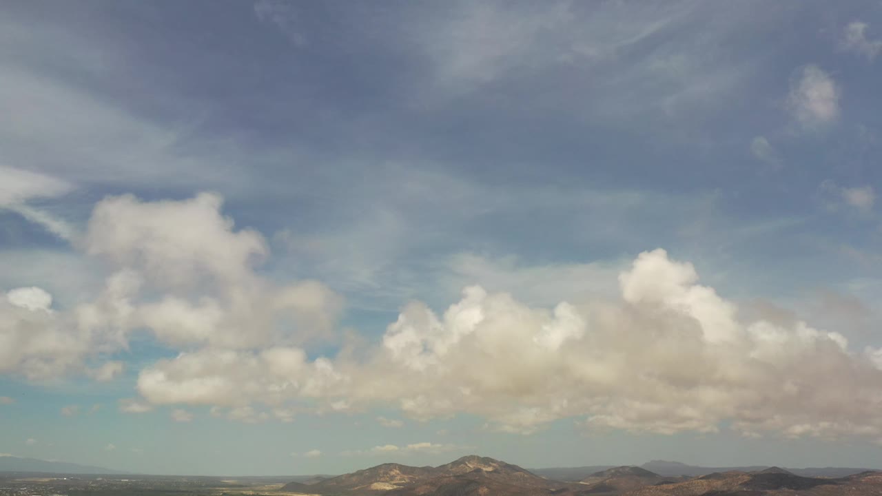Los Cabos aerial frame showing a band of valley terrain and rooftops below low brown hills, with broad blue sky and white cloud masses above, and no visible people.