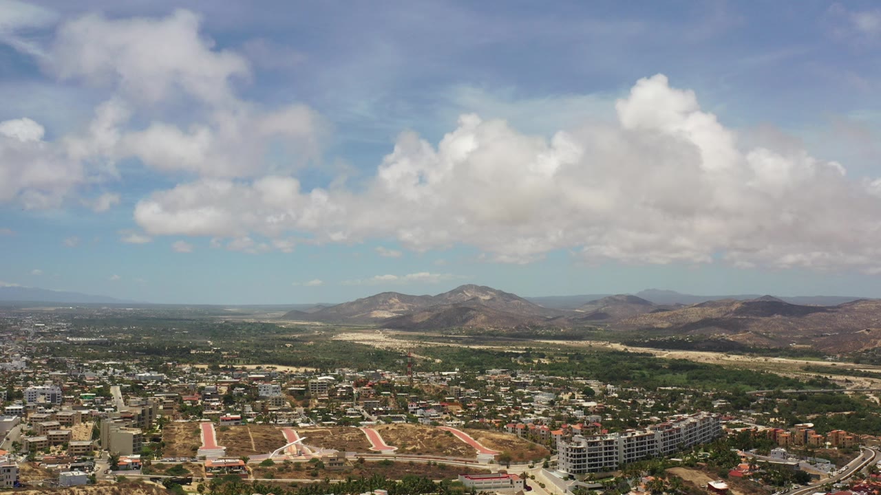 Opening aerial frame over Los Cabos, Mexico, showing city blocks in the foreground, dry open land, distant mountains, large white clouds, and no visible people.