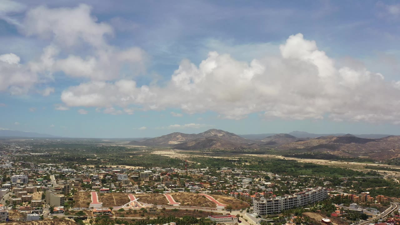Second aerial frame over Los Cabos, Mexico, with streets and low-rise buildings below, open dry lots at center, mountains beyond, and no visible people.