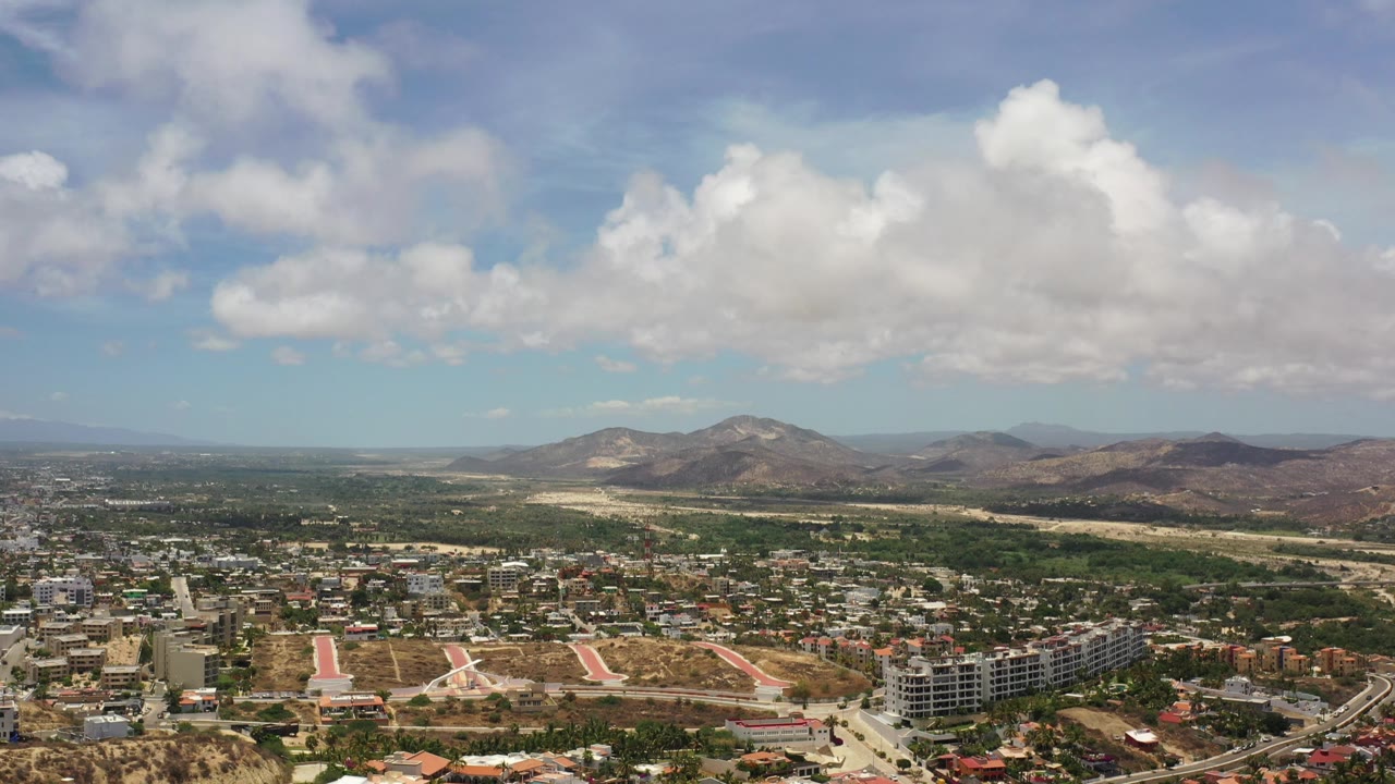 High city overview with hotel-like buildings, roads, hills, mountains, and cloud cover