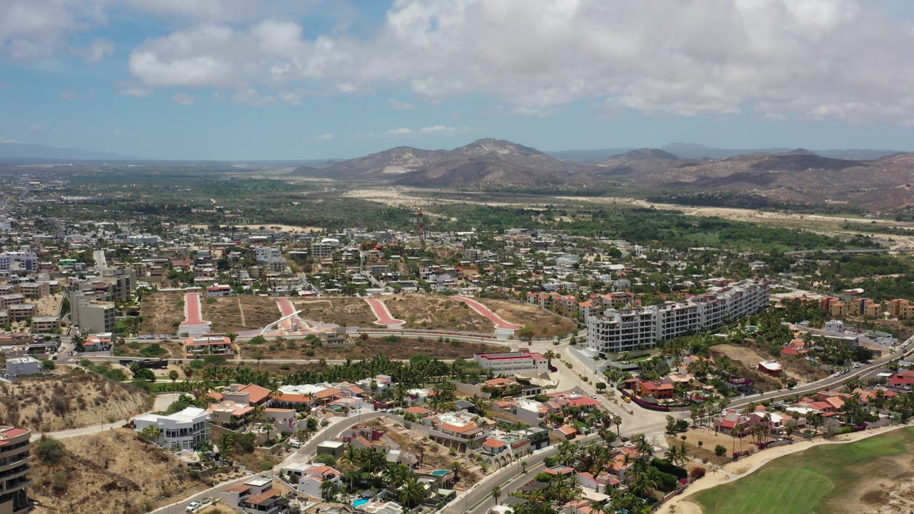 Aerial view of city roads, buildings, green trees, and dry mountains under clouds.
