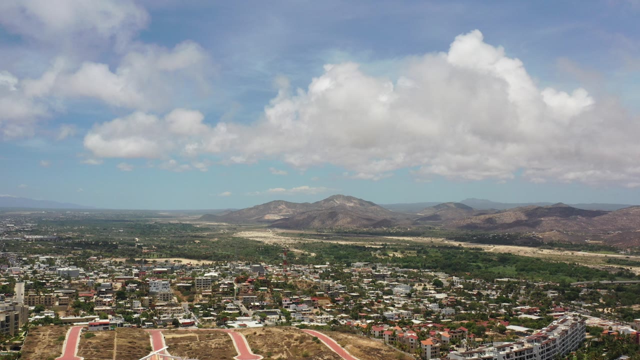 Opening frame of Los Cabos, Mexico, seen from above with low-rise buildings at the bottom, dry terrain and mountains beyond, large white clouds overhead, and no visible people.