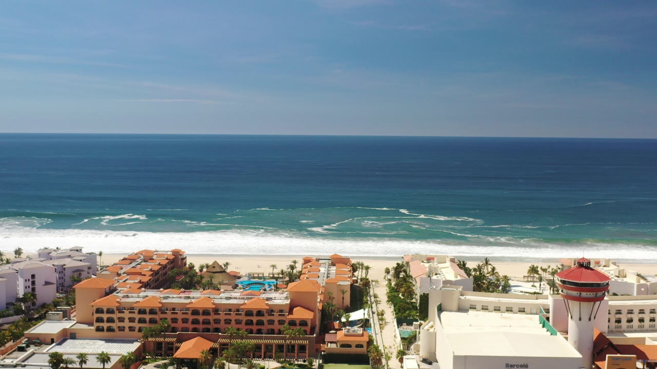 Wide aerial frame over Los Cabos, Mexico showing blue ocean, white surf, a sandy beach, orange-roof resort buildings, pools, palms, and no visible people.