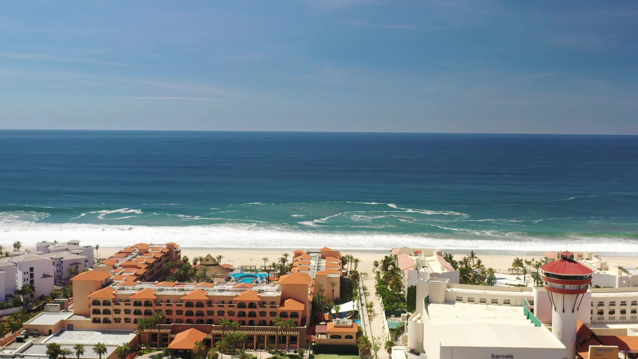 A Los Cabos aerial frame shows beachfront resort rooftops and turquoise pools below a bright surf line, with the ocean horizon high in frame and no visible people.
