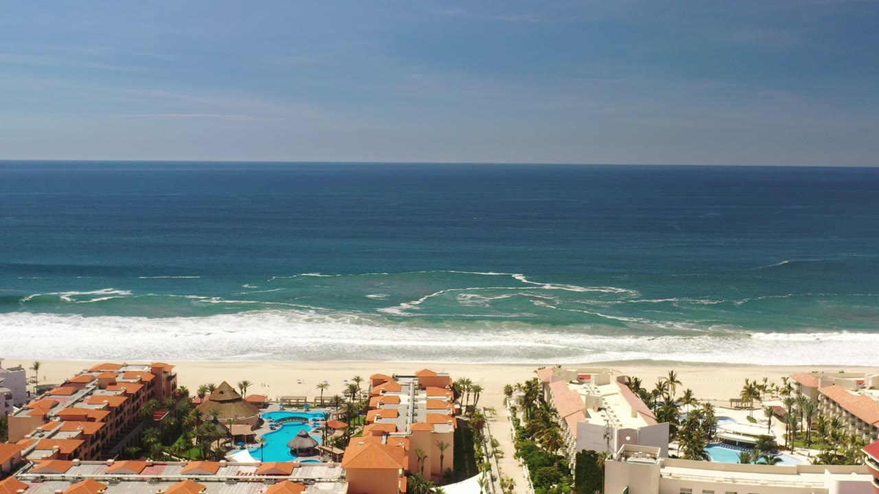 Opening aerial frame over Los Cabos, Mexico, showing blue ocean, white surf, sandy beach, orange-roofed townhomes, palms, and pool areas, with no visible people.