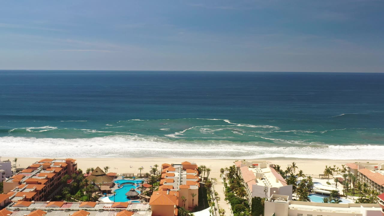Aerial frame in Los Cabos, Mexico, with the surf line slightly farther across the beach and blue pools visible among beachfront homes, with no visible people.
