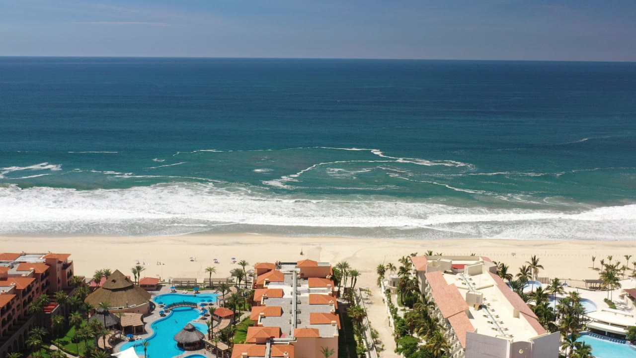 High aerial frame over a Los Cabos beach, with blue ocean and white surf beyond pale sand, resort rooftops and pools along the lower edge, and no visible people.
