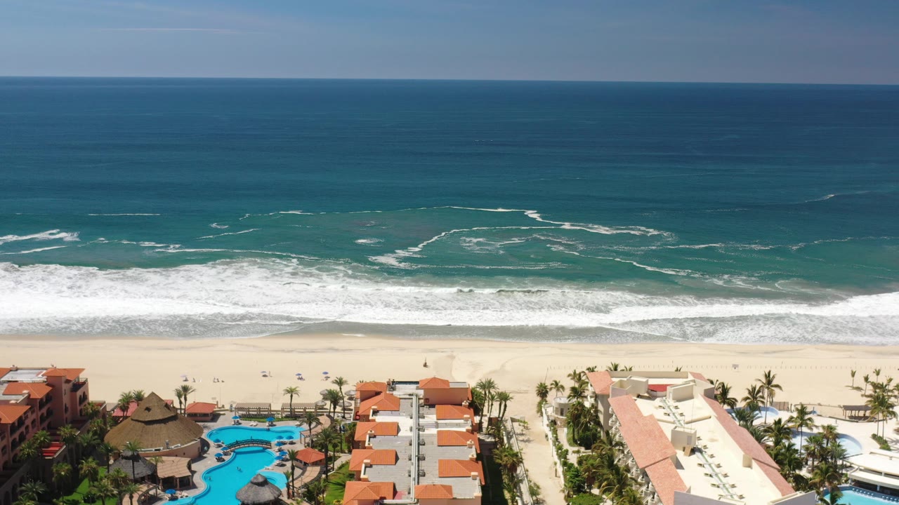 Drone-height beach view with turquoise shallows, surf, sand, palms, and pools.