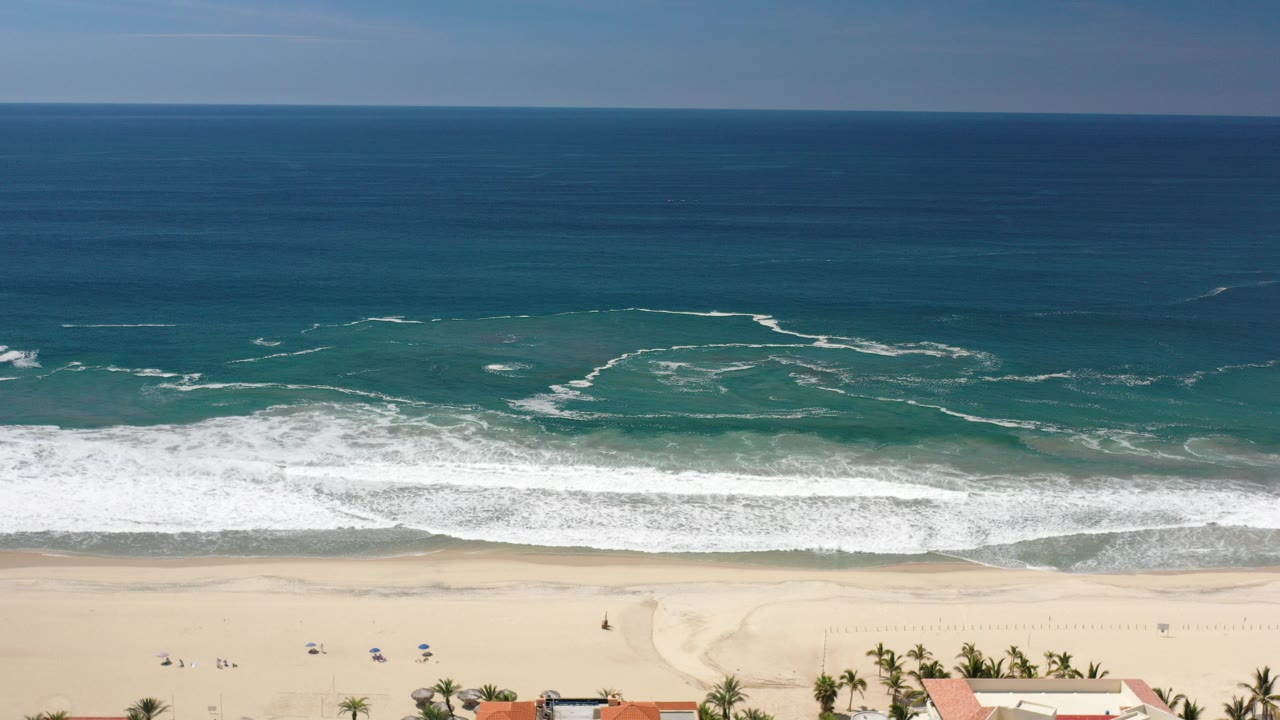 Aerial view over Los Cabos, Mexico with deep blue ocean, white surf lines, pale sand, and tiny beachgoers near umbrellas at the lower edge.