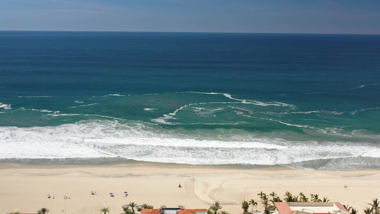 Wide aerial frame in Los Cabos, Mexico showing layered blue water, curling white foam, pale beach sand, and very small beachgoers near the shoreline.