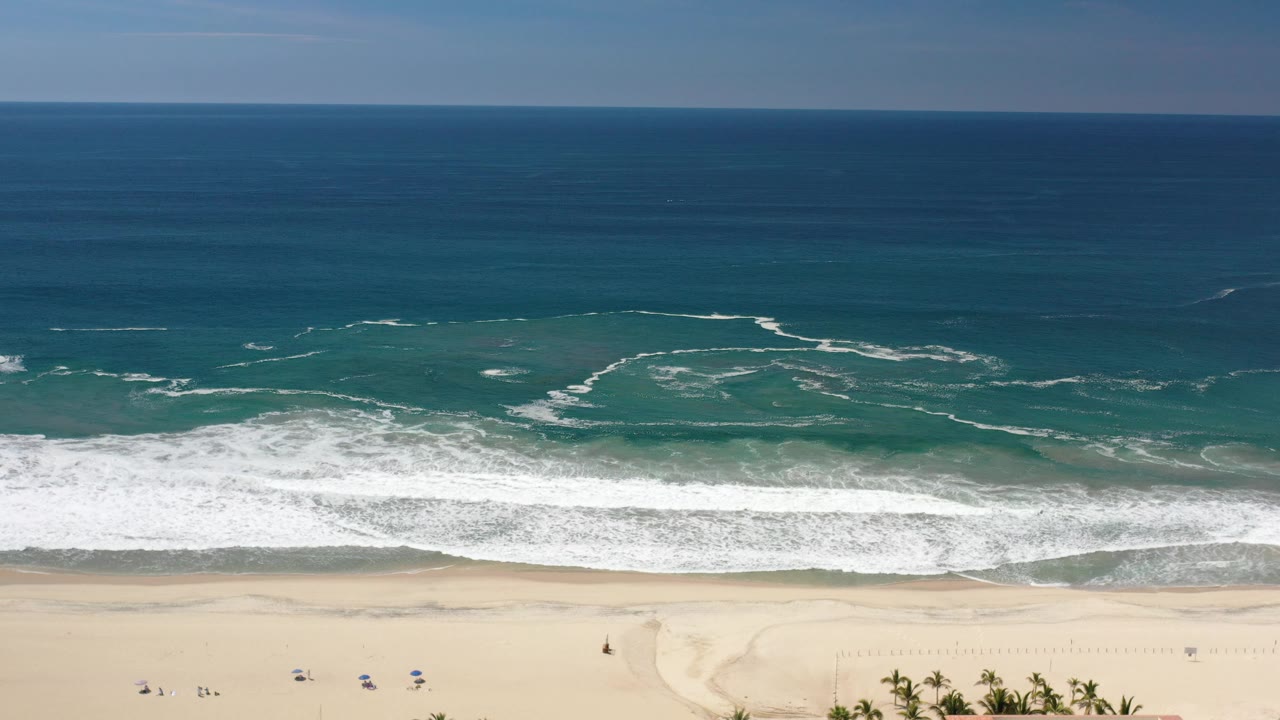 Overhead Los Cabos, Mexico beach view with white foam arcs across green-blue water, a bright sand strip, and small figures spread along the beach.