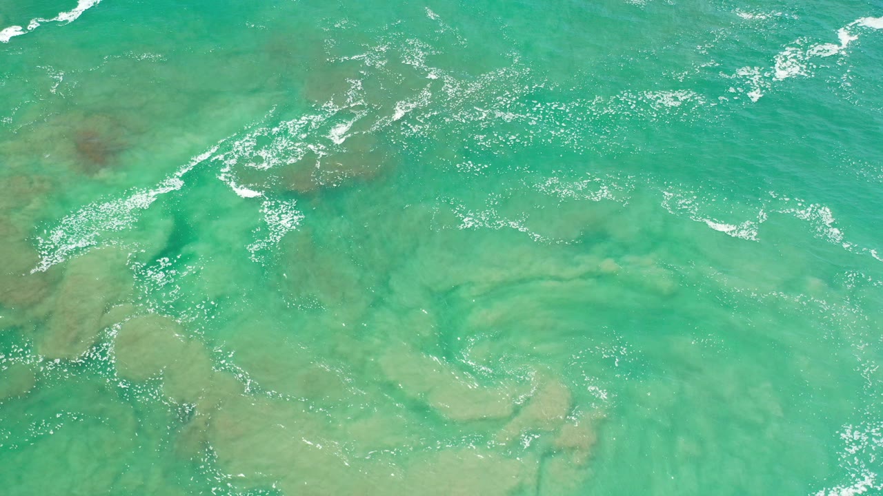 Overhead view of transparent turquoise sea water in Los Cabos, Mexico, with pale submerged rock shapes, scattered foam, and no visible people.