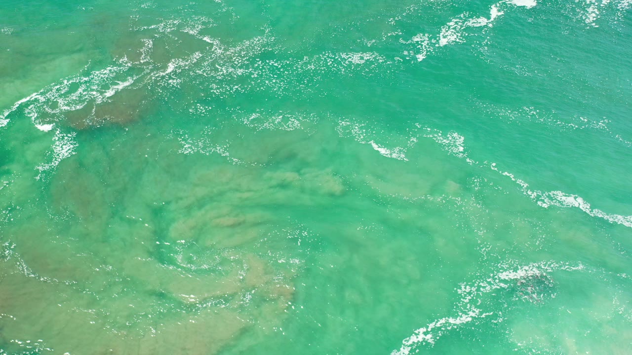 Overhead frame from Los Cabos, Mexico showing green turquoise shallow water, submerged sand or rock patches, white foam streaks near the top edge, and no visible people.