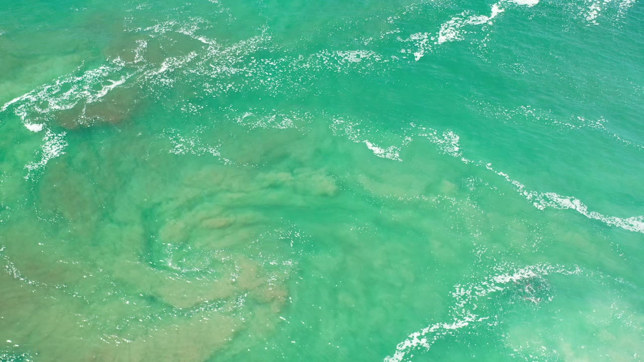 Aerial view of shallow green ocean with foam near darker submerged patches