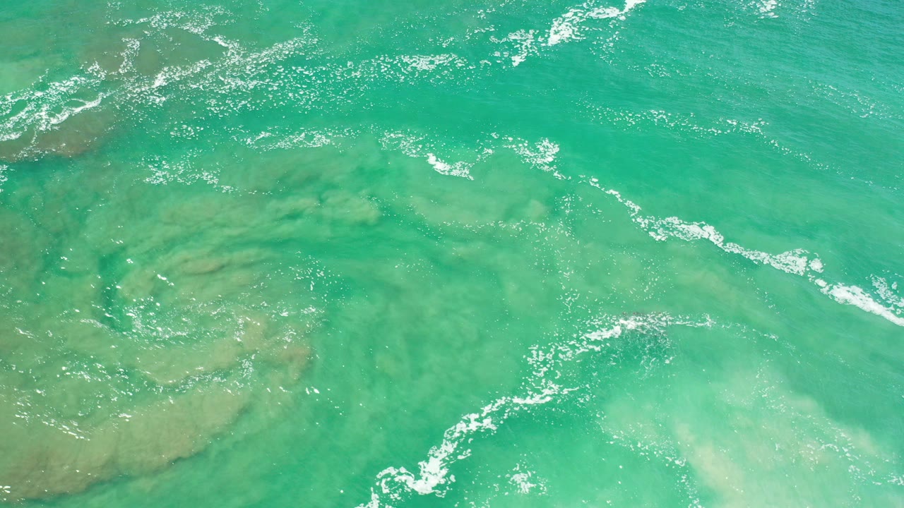 Overhead view of turquoise coastal water in Los Cabos, Mexico, with thin white foam streaks and pale submerged patches, no visible people.