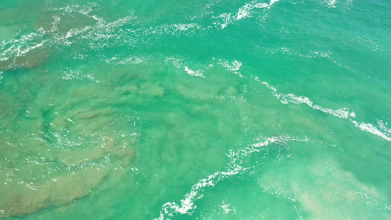 Overhead Los Cabos water frame with diagonal white foam, green-blue surface color, and cloudy shallow areas, no visible people.