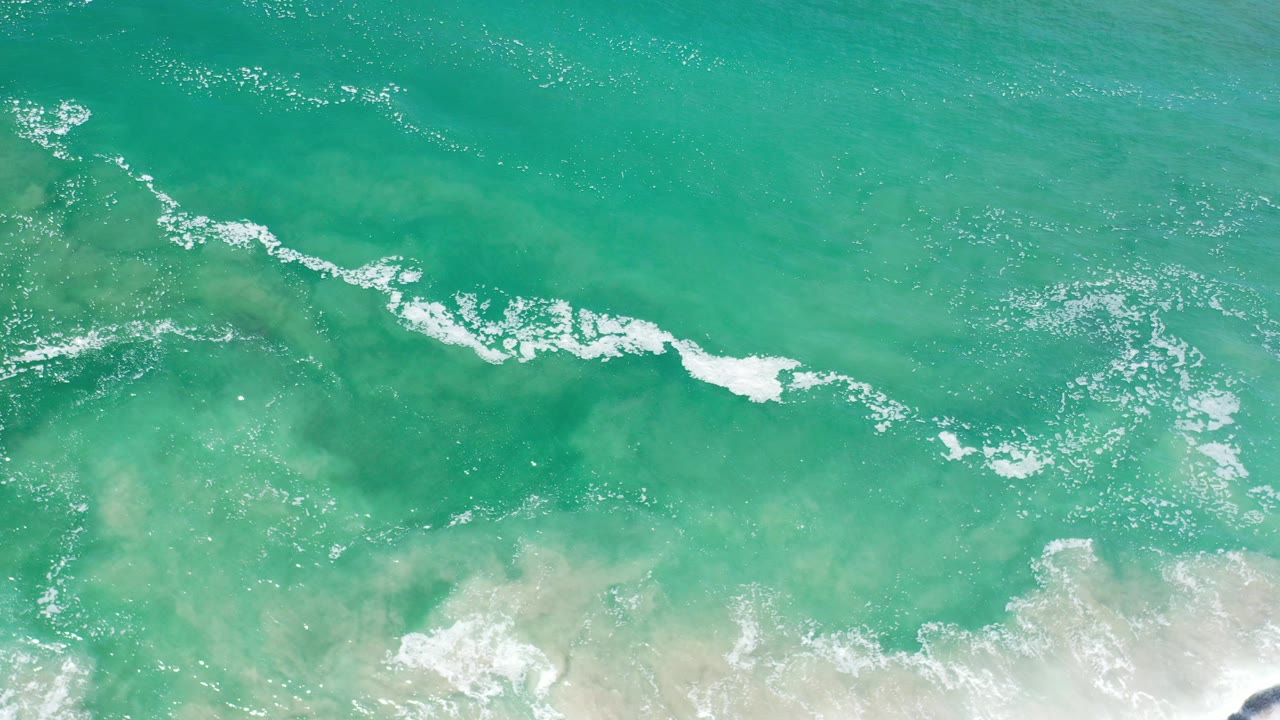 Overhead view of turquoise ocean water in Los Cabos, Mexico, with a white foam line crossing the shallow surf and pale sand at the lower edge, no visible people.