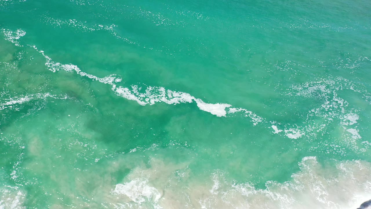 Overhead Los Cabos water view with a broken white wave line drifting across green-blue shallows, darker submerged patches, and no visible people.