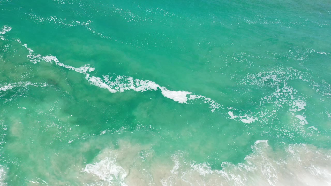 Turquoise surf seen from above with white foam and sandy shallows.