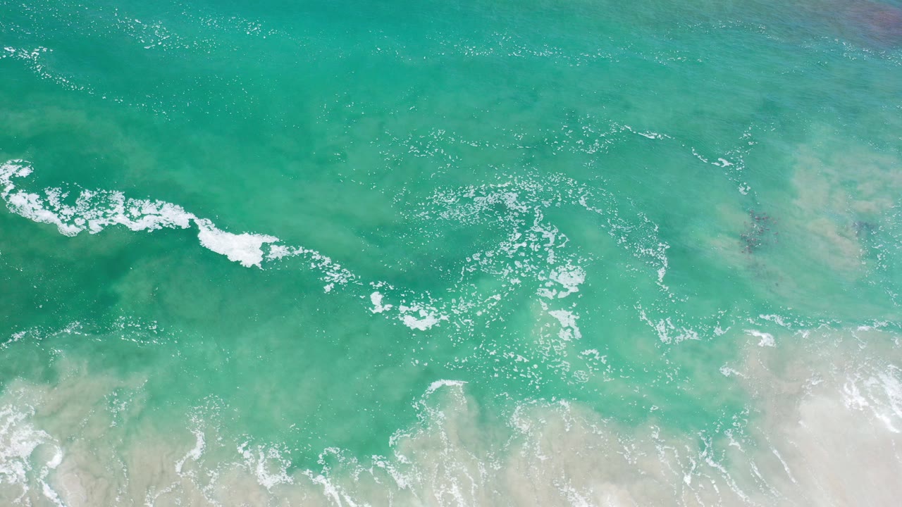 Overhead view of turquoise water and white foam crossing pale shoreline sand in Los Cabos, Mexico, with no visible people.