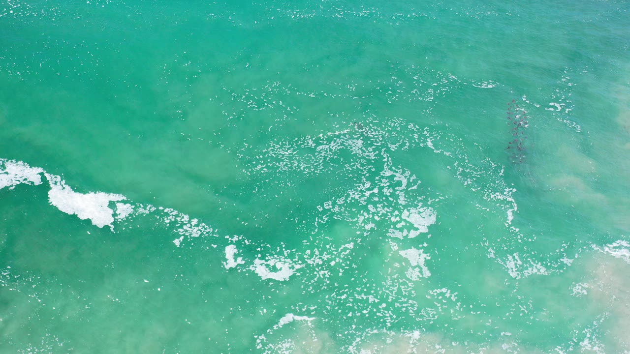 Overhead view of teal ocean water in Los Cabos, Mexico, with scattered white foam near the left side, pale underwater shallows, and no visible people.