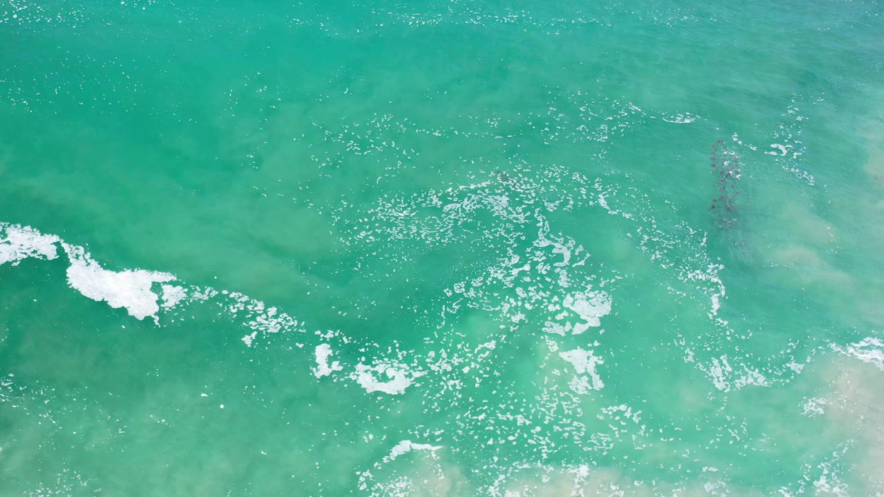 Aerial frame over Los Cabos teal ocean, showing a brighter white foam streak at left, rippled water across the center, pale shallows, and no visible people.