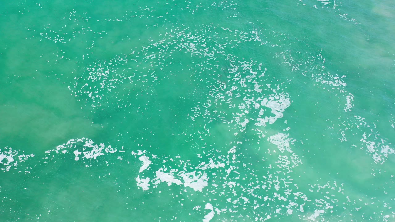 Top-down view of turquoise ocean water in Los Cabos with white foam patches near the lower left and no visible people.