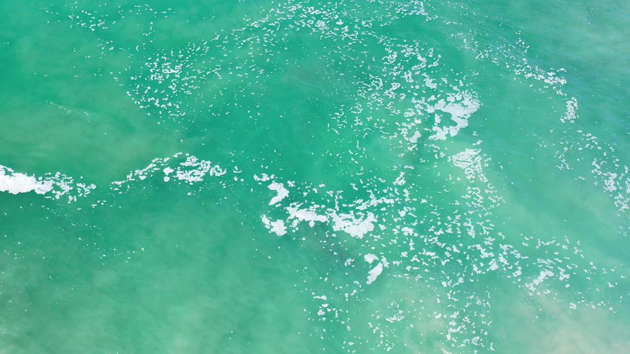 Overhead view of turquoise-green sea in Los Cabos, Mexico, with scattered white foam, bright surface sparkle, pale underwater patches, and no visible people.