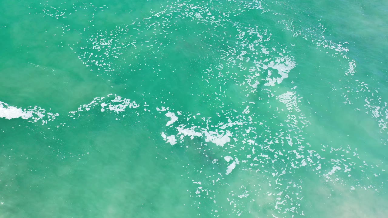Overhead Los Cabos sea surface with a narrow white foam line crossing the lower left area, sunlit green water, faint brown patches, and no visible people.