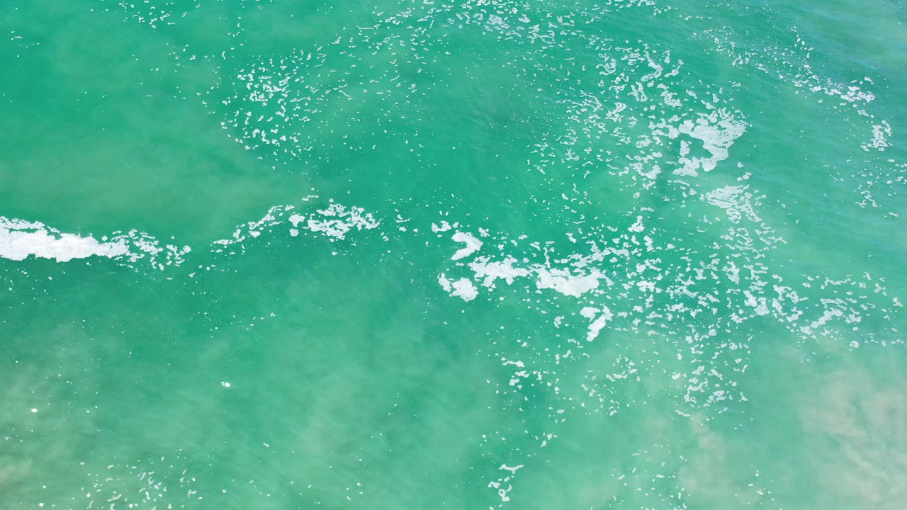 Overhead view of turquoise shallow surf and white foam over pale sand in Los Cabos, Mexico, with no visible people.