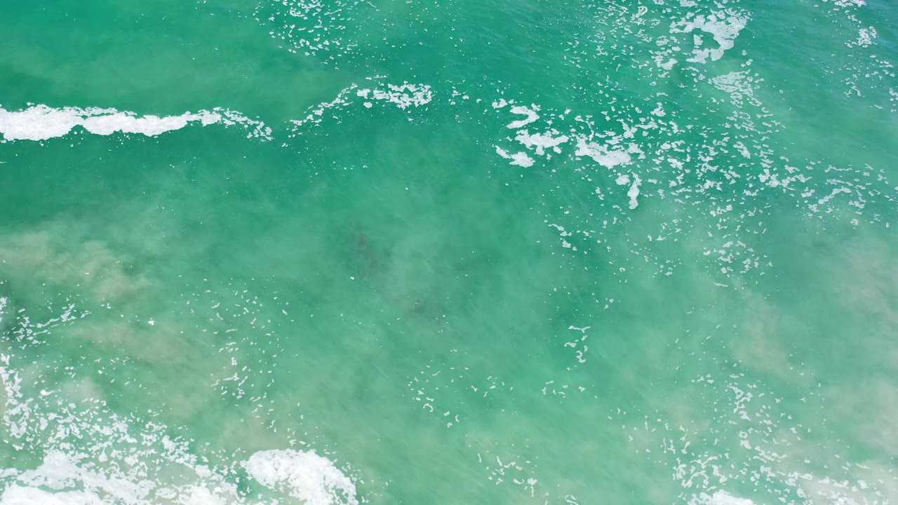 Overhead Los Cabos ocean frame showing green-blue water, a horizontal foam streak near the top, pale sand below the surface, and no visible people.