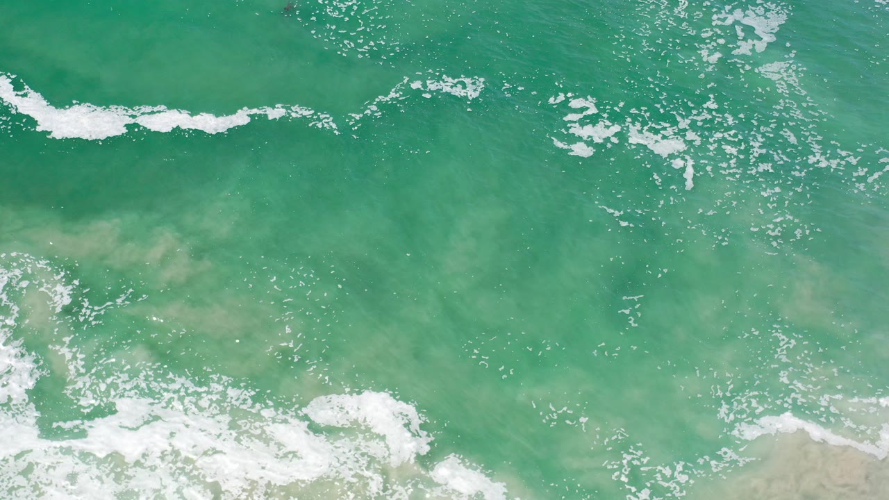 Top-down view of green surf and white foam over sandy shallows in Los Cabos, Mexico, with no visible people.