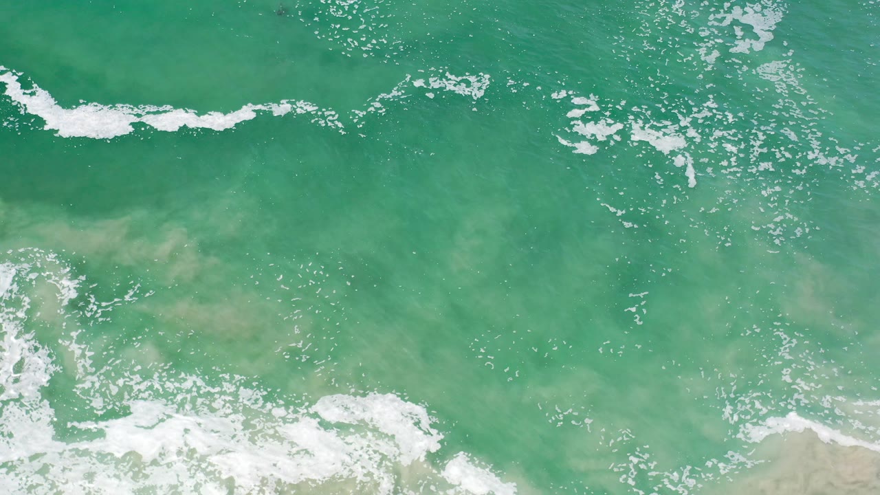Overhead turquoise surf in Los Cabos, Mexico shows foam breaking near pale sandy shallows, with no visible people.