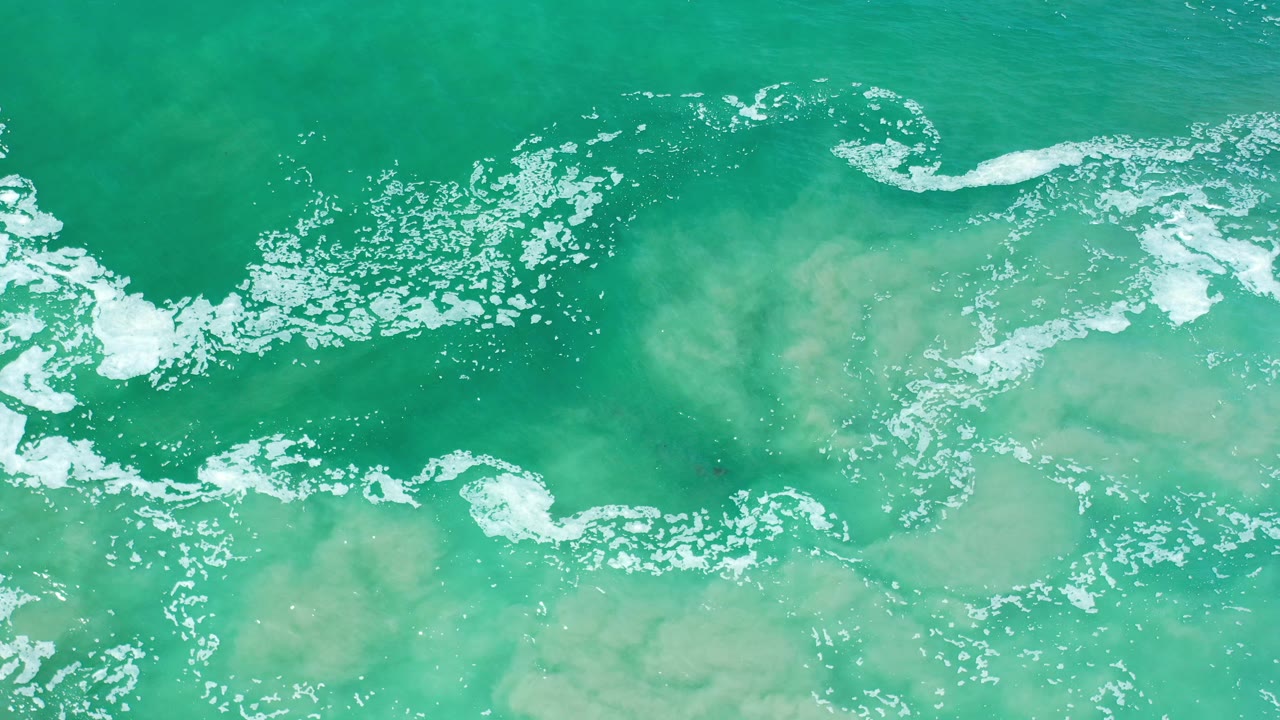 Overhead view of aqua green ocean water in Los Cabos, Mexico, with scattered white foam streaks and cloudy murky patches beneath the surface, no visible people.