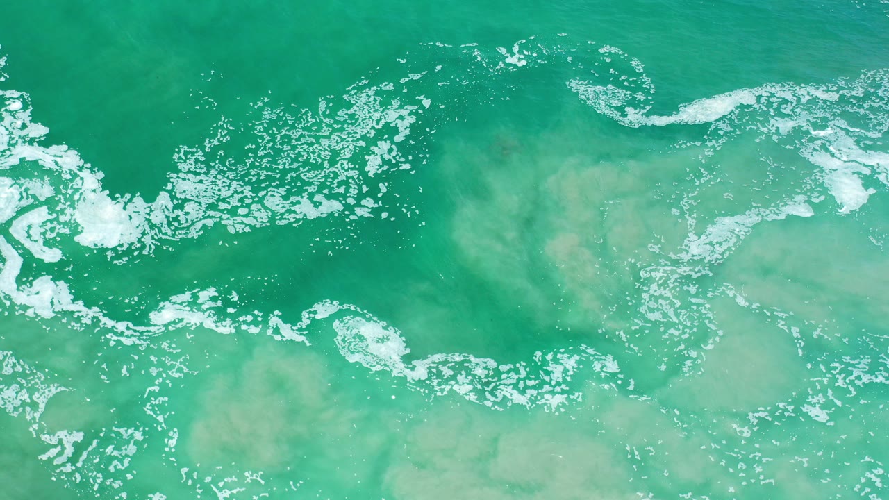 Overhead view of turquoise ocean water in Los Cabos, Mexico, with white foam crossing the upper frame and no visible people.