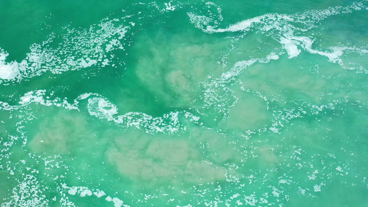 Overhead view of green Los Cabos seawater with white foam bands near the top and pale sandy churn under the surface, with no visible people.