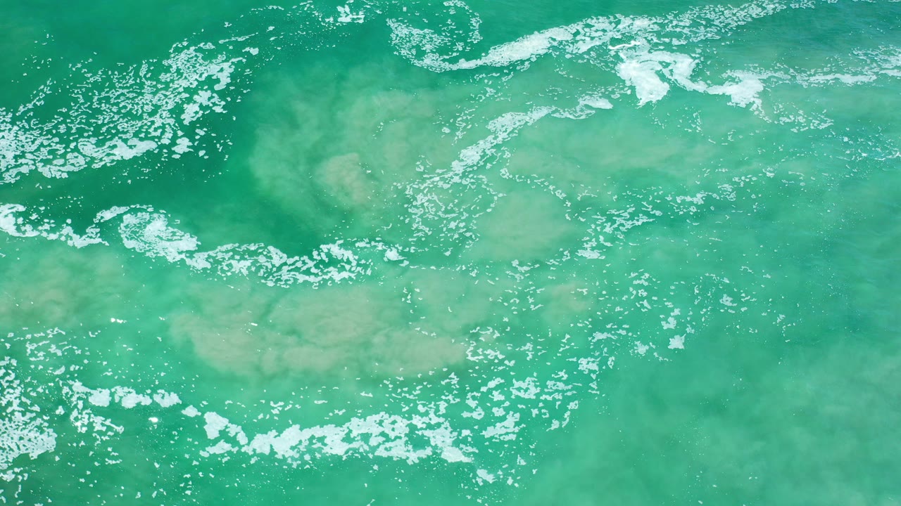 Opening overhead view of green seawater in Los Cabos, Mexico with a pale submerged sand patch near center and white foam scattered across the surface, no visible people.