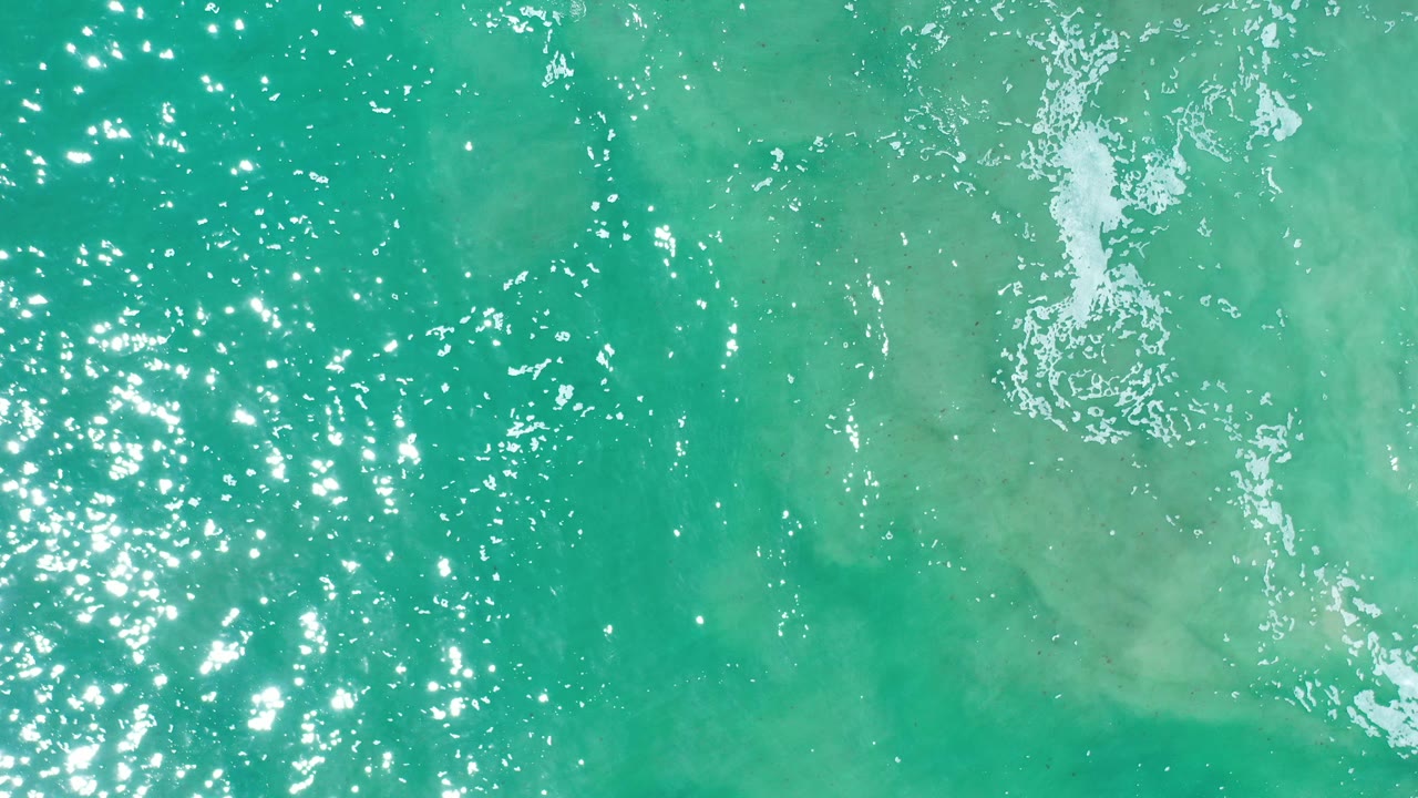 Overhead view of turquoise-green ocean water in Los Cabos, Mexico, with bright sun reflections, rippled texture, and a narrow patch of white froth near the upper right, with no visible people.