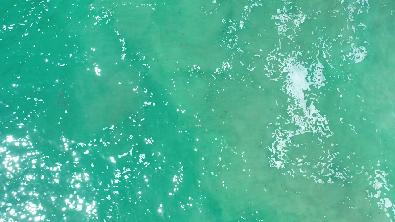 Overhead view of turquoise sea water in Los Cabos, Mexico, with scattered white foam near the upper frame and no visible people.