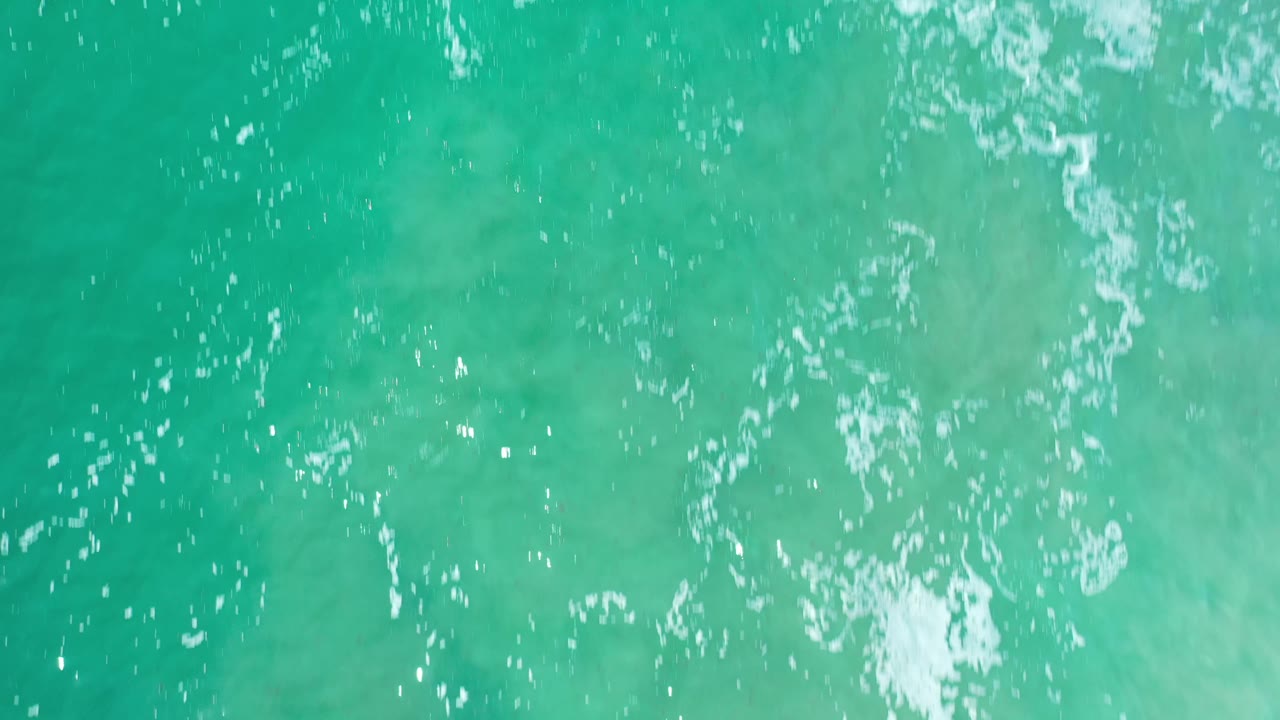 Clear green-blue water in Los Cabos, Mexico, seen from above with scattered white reflections and a thin foam streak near the top, with no visible people.