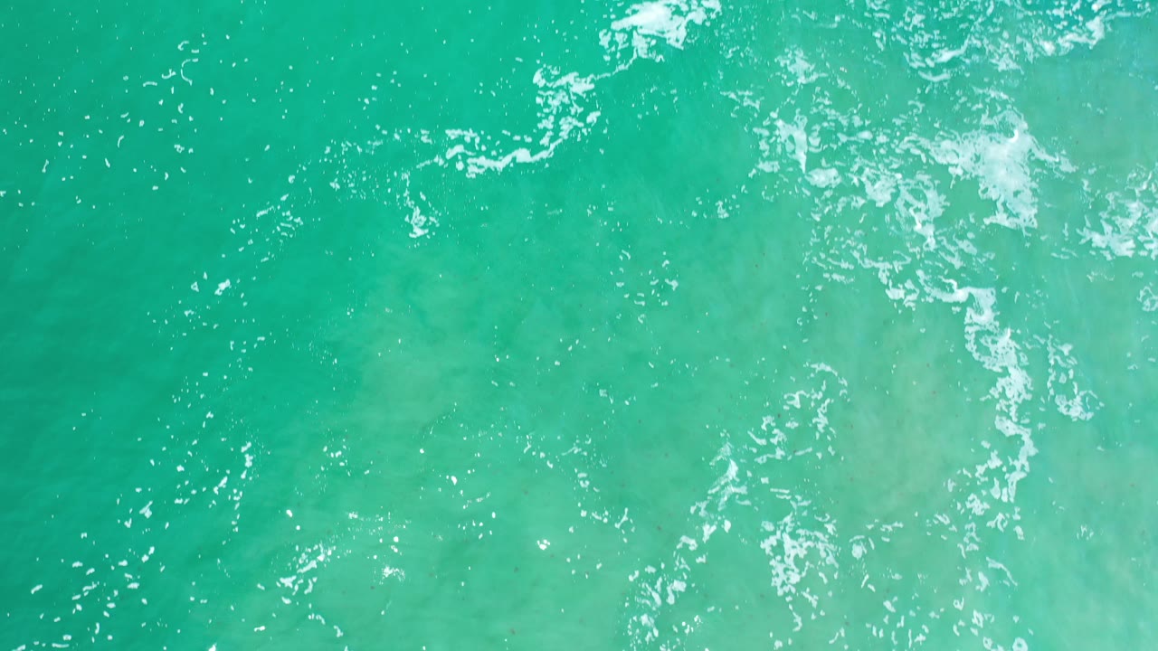 Overhead ocean surface with white foam and pale shallows.