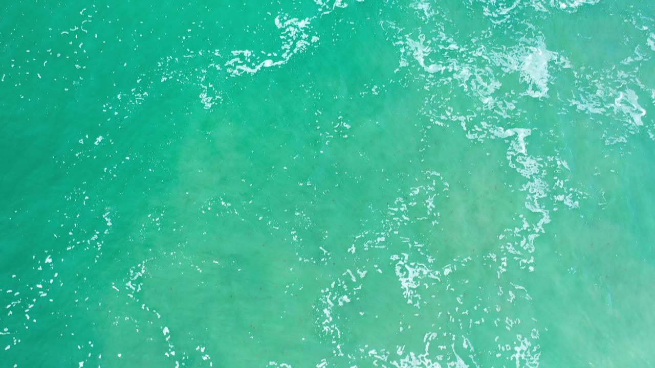 Overhead view of turquoise-green sea water in Los Cabos, Mexico, with scattered white foam near the top of frame and no visible people.