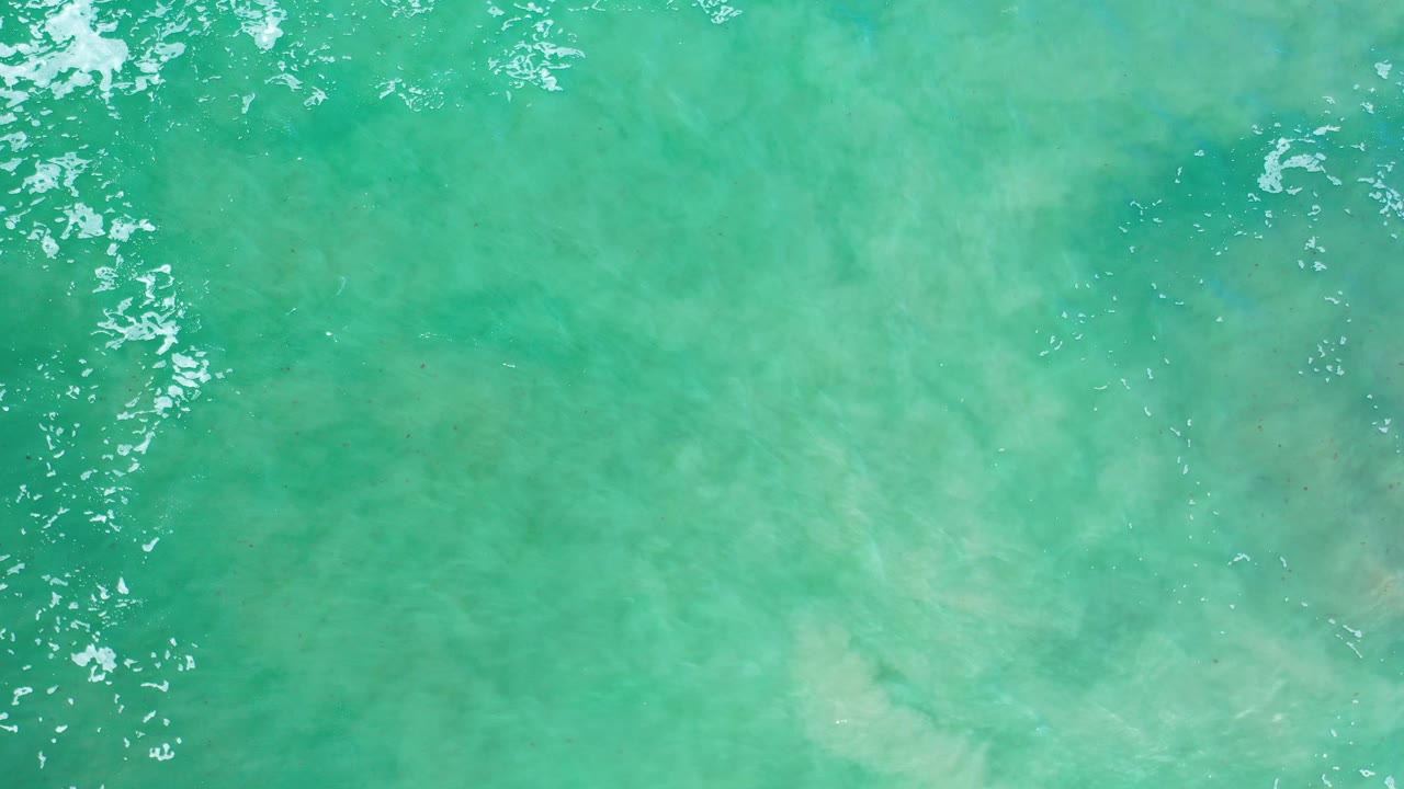 Overhead view of turquoise ocean water in Los Cabos, Mexico, with pale submerged texture and scattered white foam flecks near the top edge, no visible people.