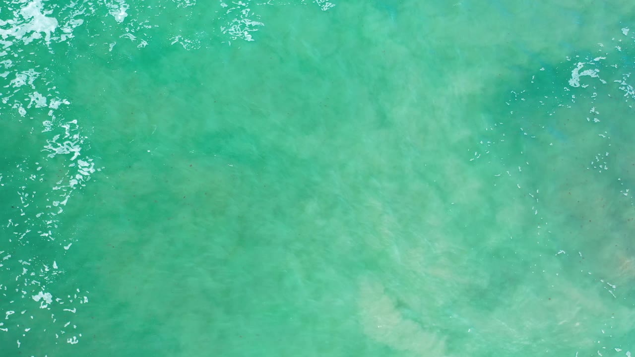 Blue-green ocean surface in Los Cabos, Mexico, with brighter sun reflections along the upper water and pale sand-like patches beneath the surface, no visible people.